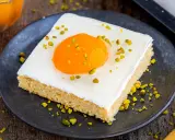 Ein Stück Kuchen mit weißer Glasur, einem orangen Gelee-Dotter und gehackten Pistazien, das wie ein Spiegeleikuchen mit Quark aussieht.