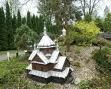 Drewniana cerkiewka z metalowym dachem, miniatura w parku miniatur Myczkowce, otoczona zielenią i kamieniami.