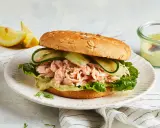 Ein Brötchen mit pulled Lachs, Salat, Gurkenscheiben und einer cremigen Sauce. Dazu Zitronenspalten und ein Dip.