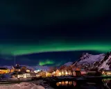 Lofoten norrsken över en snötäckt by med upplysta hus och speglingar i vattnet.