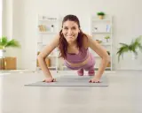 Frau in lila Sportkleidung macht Liegestütze für Yoga Muskelaufbau. Sie lächelt glücklich in die Kamera.