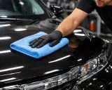 Mężczyzna w czarnej rękawiczce poleruje czarny lakier auta niebieską mikrofibrą. Czy po woskowaniu płukać auto? To pytanie zadaje sobie wielu kierowców.