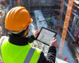 Ingeniero con casco naranja y chaleco reflectante revisa planos en tablet. Asegura el control de calidad en construcción.