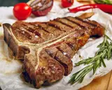 Saftiges T-Bone-Steak, perfekt gegrillt, mit grobem Salz bestreut, daneben Rosmarin, Tomaten und Chili.