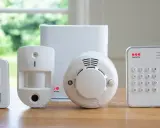 En uppsättning säkerhetskomponenter: en rörelsedetektor, en dörr-/fönstersensor, en rökdetektor och ett kontrollpanel med knappsats. Allt redo för att ge en alert alarm.