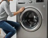 Kobieta zamyka drzwi nowoczesnej, srebrnej pralki LG. Obok stoi drewniany regał z ręcznikami.