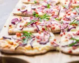Flammkuchen-Ideen: Dünner Teig mit Käse, roten Zwiebeln, Speck und Rucola belegt. Perfekt für den Sommer!