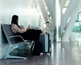 Kobieta czeka na lotnisku, obok niej stoi szary sas bagaż rejestrowany i plecak.