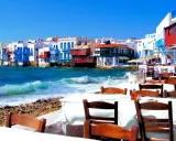 Cosa vedere a Mykonos in crociera: case colorate sul mare, onde che si infrangono sulla spiaggia di ciottoli e tavoli apparecchiati per godersi il panorama.