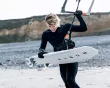 Młody kitesurfer w czarnym piance, doceniając jej właściwości termoizolacyjne, przygotowuje deskę do wodnych szaleństw.