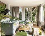 Sofá gris y otomanas verdes crean un ambiente acogedor. Plantas exuberantes y luz natural inundan el salón.