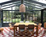 Salle à manger lumineuse dans une véranda avec toiture zinc. Table en bois, chaises colorées, sol à damier rouge et blanc, vue sur jardin verdoyant.