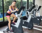 Uśmiechnięta kobieta na rowerku stacjonarnym. Ćwiczenia na rowerku angażują mięśnie nóg i pośladków.