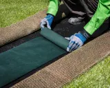 Manos enguantadas desenrollan césped artificial verde sobre una base negra. Paso clave para saber como colocar cesped artificial.