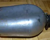 Metalowa manierka US Army z czarną zakrętką i łańcuszkiem. Widoczne ślady użytkowania i rdzy.