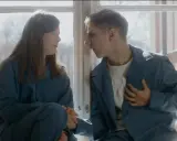 Två unga människor, klädda i blåa arbetskläder, sitter vid ett fönster. Scen från filmen om vipeholmsexperimenten.
