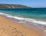 Türkisblaues Meer und ein sandiger Strand bei Oran Stadt. Wellen brechen sanft, im Wasser sind einige Schwimmer zu sehen.