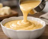 Une louche verse une sauce onctueuse et dorée dans un bol blanc. C'est la sauce au langres, prête à être dégustée.
