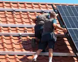Två personer arbetar med att montera solceller på ett tegeltak. De fäster solpanelerna på monteringsskenor.