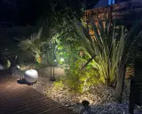 Jardin illuminé la nuit, créant une ambiance chaleureuse. La **norme éclairage extérieur** est respectée pour mettre en valeur la végétation luxuriante et le chemin en bois.