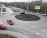 Rowerzysta na rondzie, otoczony przez czerwone samochody i drzewa.