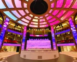 Interni di una nave da crociera, con un palco illuminato da luci rosa e blu, che ricorda la costa mediterranea.