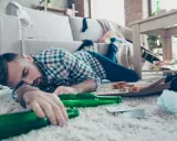 Mężczyzna śpi na dywanie, otoczony pustymi butelkami i resztkami jedzenia. To obrazek pokazujący skutki nadużywania alkoholu, jak padaczka alkoholowa po jakim czasie picia może się pojawić.