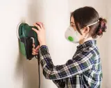 Mujer con mascarilla lijando una pared con una lijadora eléctrica.