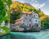 Antica casa in pietra con edera su un lago turchese, un ponte ad arco e colline verdi sullo sfondo, un angolo da fiaba dei castelli Lombardia.