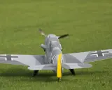 Modellflugzeug einer Me109 mit Tarnmuster und gelbem Seitenleitwerk auf einer grünen Wiese.