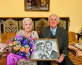 Para świętuje 70 rocznicę ślubu, trzymając zdjęcie z dnia zaślubin. W bukiecie kwiatów dominują fiolety i pomarańcze.