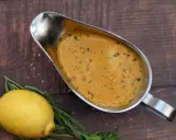 Eine cremige Cafe de Paris Sauce in einer Sauciere, daneben ein Zitrone und Kräuter auf dunklem Holz.