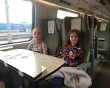 Dzieci siedzą w przedziale dla osoby z dzieckiem do lat 6 Intercity. Chłopiec w kolorowym T-shircie i dziewczynka w kurtce z kwiatowym wzorem.