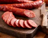 Salami auf einem Holzbrett, geschnitten und im Ganzen. Man kann Salami einfrieren, um sie länger haltbar zu machen. Knoblauch und Messer liegen daneben.