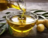 Aceite de oliva virgen extra vertiéndose en un cuenco de cristal, rodeado de aceitunas y hojas de olivo sobre madera rústica.