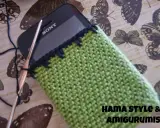 Funda de móvil tejida a crochet, un patrón verde con borde negro. Un gancho y hilo negro están listos para continuar el proyecto.