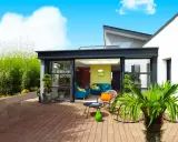 Une véranda moderne avec de grandes baies vitrées s'ouvrant sur une terrasse en bois. Un salon coloré est visible à l'intérieur.