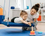 Terapia integracji sensorycznej: dziewczynka na huśtawce z terapeutką, która pomaga jej sięgnąć po zabawkę.