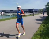 Triathlet im blauen Anzug läuft auf einer Straße. Er fragt sich, wie wird man schneller. Ein Radfahrer ist im Hintergrund zu sehen.