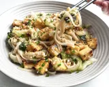 Pyszny makaron z tofu, posypany orzeszkami ziemnymi i szczypiorkiem, podany w szarej misce.