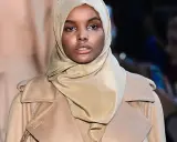 Halima Aden walks the runway in a stylish beige coat and hijab, exuding confidence and grace.