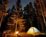 Nocne pole namiotowe nad jeziorem. Rozświetlony namiot, ognisko i ławka wśród drzew pod rozgwieżdżonym niebem.