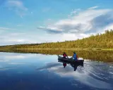 Två personer paddlar kanot på en spegelblank sjö i Sverige. Skogen längs stranden reflekteras i vattnet.