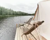 Två träfåtöljer och ett litet bord på en brygga vid en sjö, med skog i bakgrunden. En del av ett glampingtält syns. Upplev glamping Norrbotten.