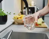Ręka nalewa wodę do szklanki. W tle widać owoce i warzywa, jakby przygotowywano posiłek. Czysta woda, jak w akwarium.