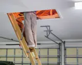 Osoba wspina się po drewnianej drabinie do włazu na strych. Widać drewniane belki stropowe i izolację. To dobry przykład, jak wykończyć właz na strych.