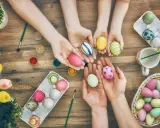 Hände von Kindern und Erwachsenen bemalen bunte Ostereier. Ein Korb voller Eier und Tulpen auf Holztisch.
