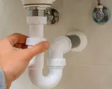 Mano ajustando la altura del desague del lavabo, tubería blanca de PVC bajo el lavamanos.