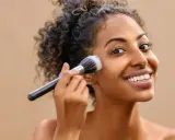Donna sorridente applica il fondotinta con un pennello. La sua pelle luminosa è il risultato di cosmetici nichel free.