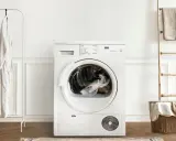 Vit torktumlare med kläder i trumman. Bredvid hänger en vit morgonrock och en beige filt. En trästege med en handduk står vid en korg med tvätt. Akryl textil i korgen.